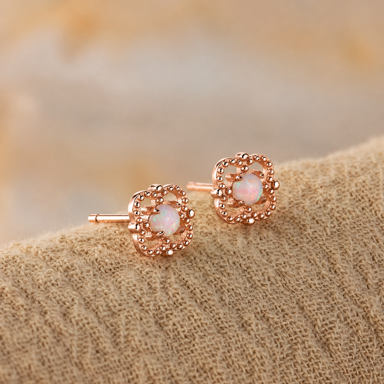 A pair of rose gold stud earrings with circular milgrain detailing and a central iridescent white opal gemstone, resting on a textured beige fabric surface.